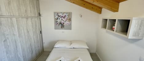 Un dormitorio con techo de madera, una cama con ropa blanca, una obra de arte floral en la pared y un estante con una botella.