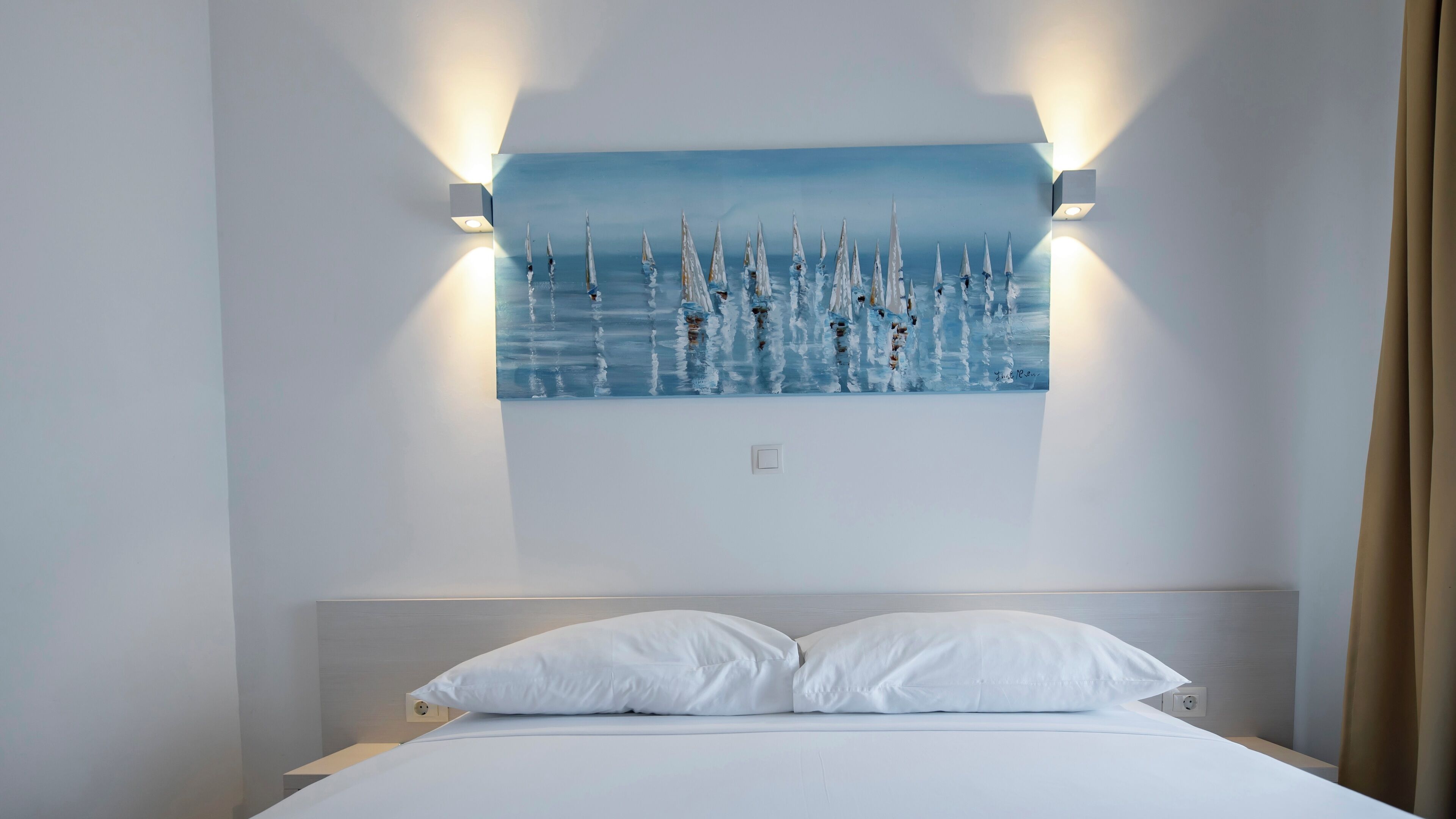 A hotel room with a bed, white pillows, a painting of a seascape, and wall-mounted lighting.