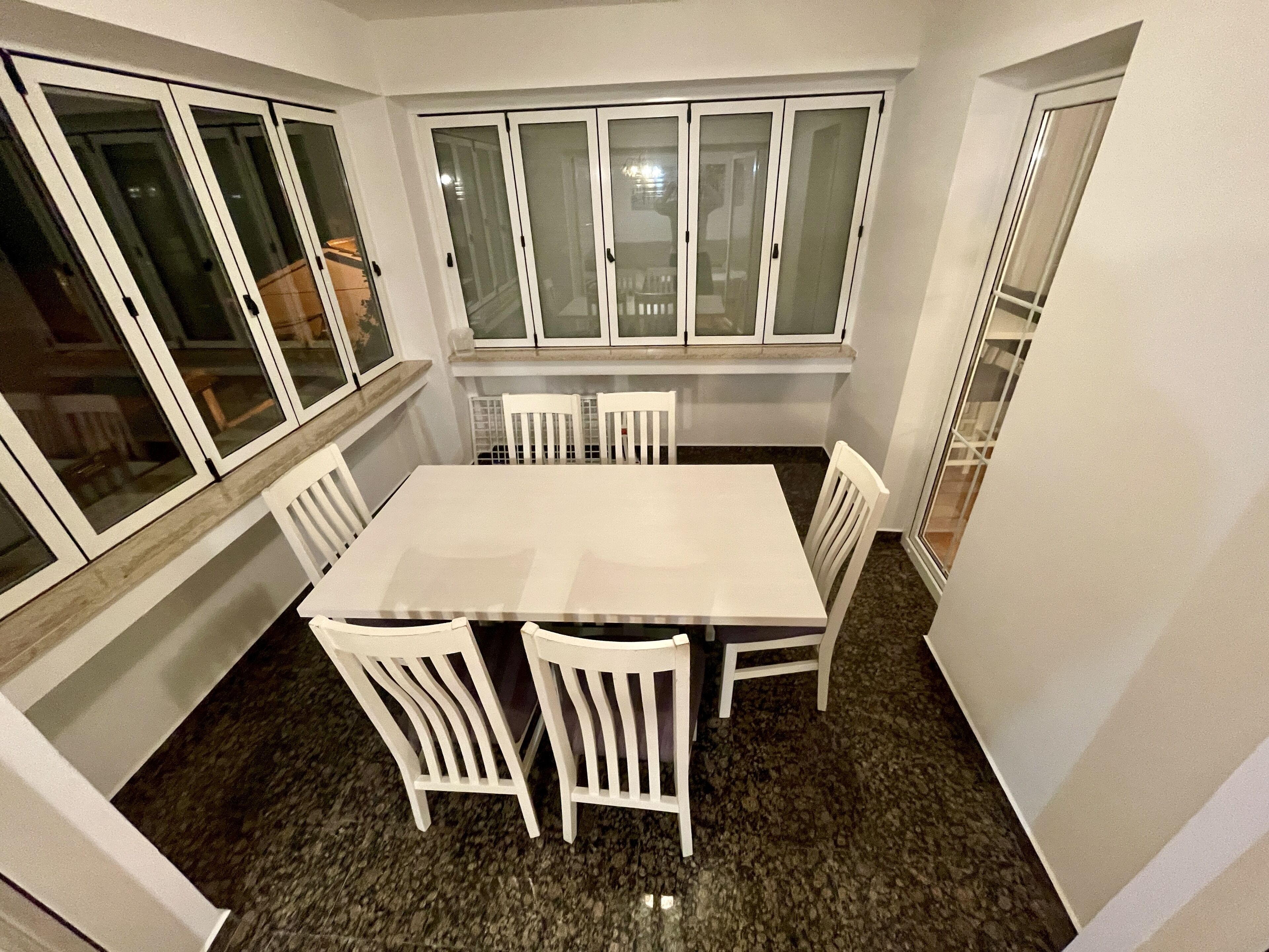 A dining area with a white table and chairs, large windows with frosted glass, and a granite floor.