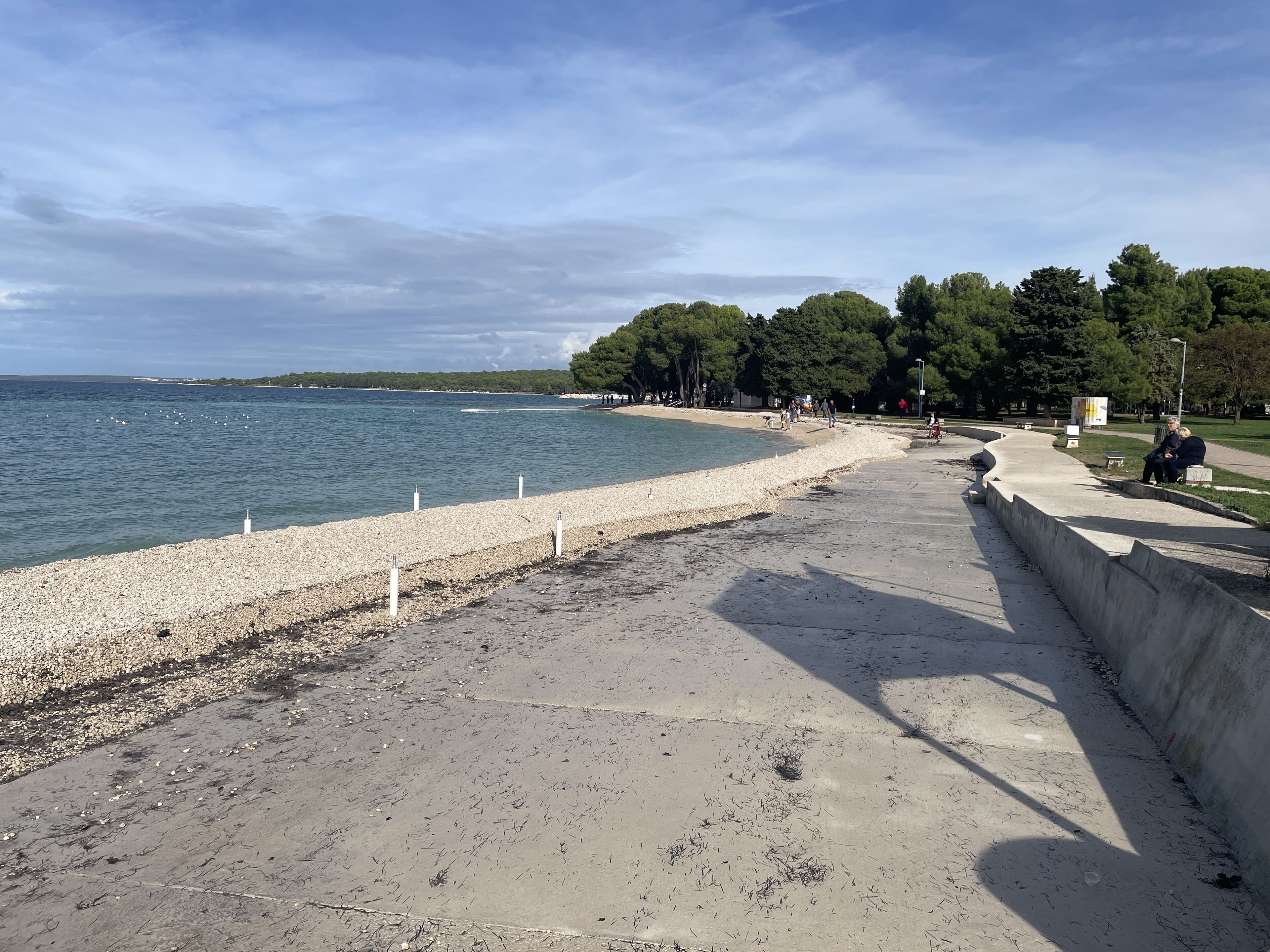 Een kustpad met een kiezelstrand, een betonnen muur en een gazon met bomen.