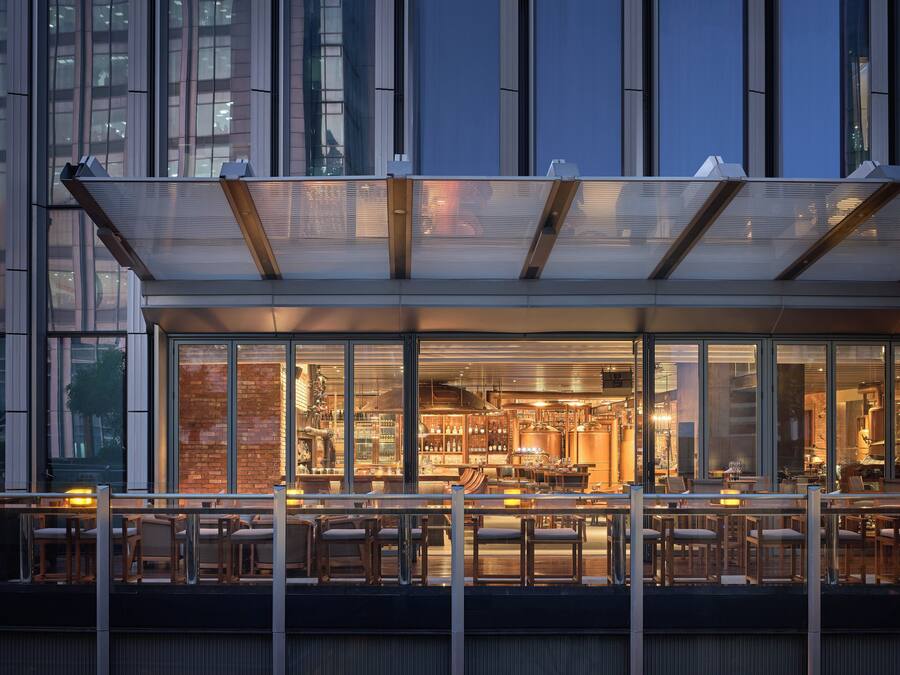 Un edificio moderno con fachada de vidrio, un bar en la azotea con mesas al aire libre y espacios interiores visibles, incluyendo una zona de cocina.