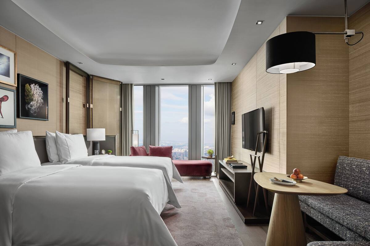 Habitación de hotel con dos camas, un sofá, una mesita, un televisor y un ventanal grande con vistas a la ciudad.