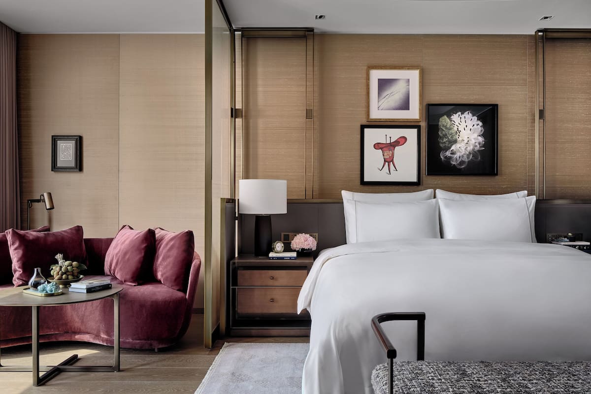 Una habitación de hotel moderna con una cama grande, un sofá rojo, una mesita de noche con lámpara y obras de arte en las paredes.