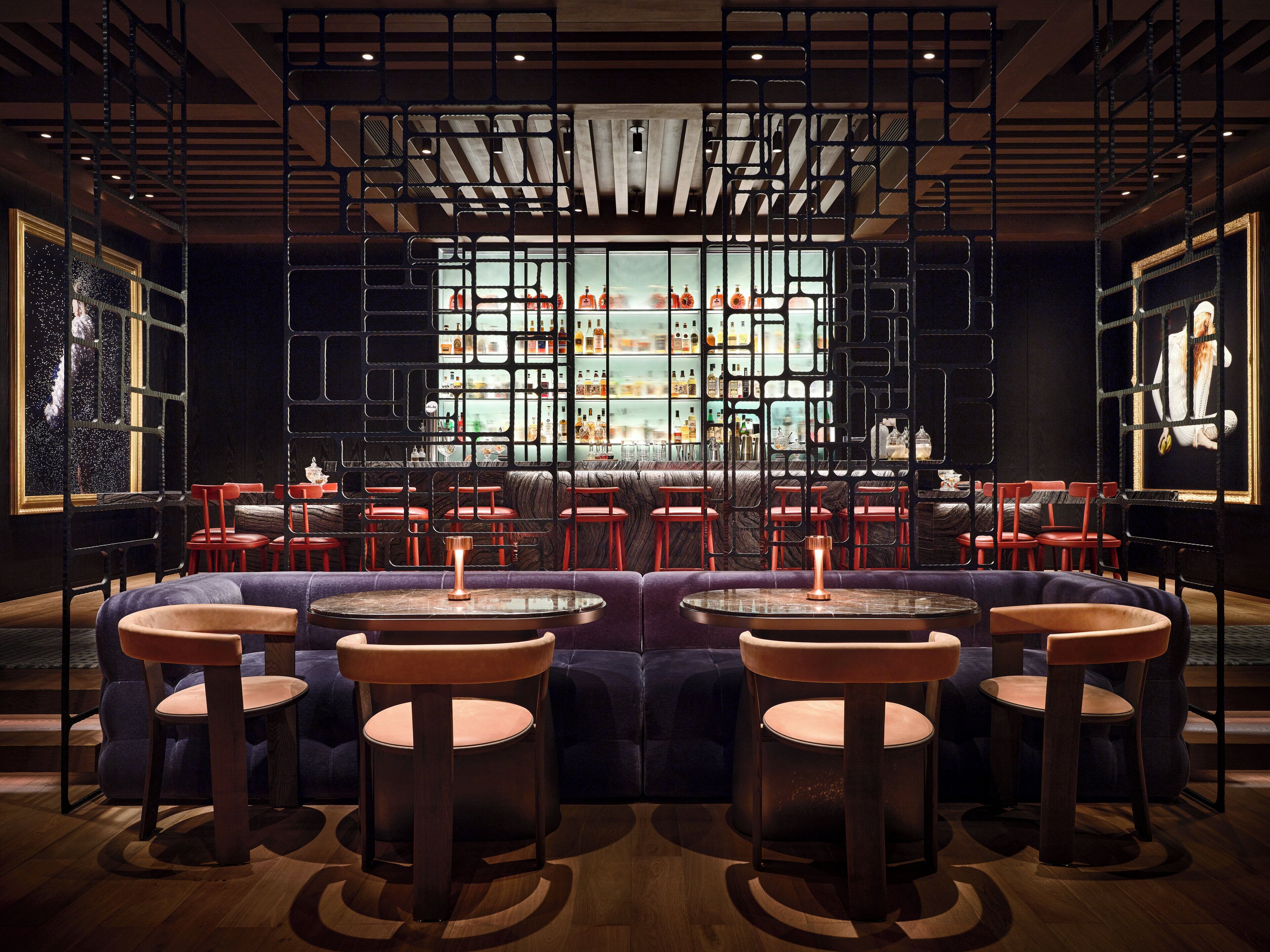 A modern bar area with round tables and red chairs, a bar counter with bottles, and a large window with a view of the city.