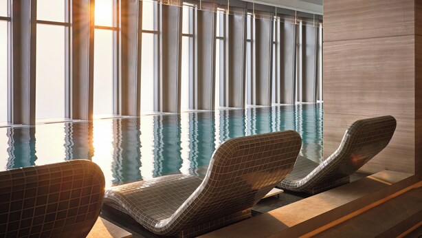 Ein Indoor-Pool mit modernem Design, der ein schlankes, längliches Becken mit klarem Wasser und Spiegelungen der Fenster zeigt. Zwei Liegestühle mit Rillenmuster sind am Poolufer platziert.