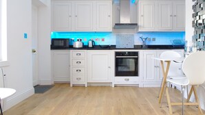 A modern kitchen with white cabinetry, a black microwave, and a built-in oven. There is a white dining table with chairs and a blue-lit backsplash. - The London Agent Chelsea Peaceful (London)