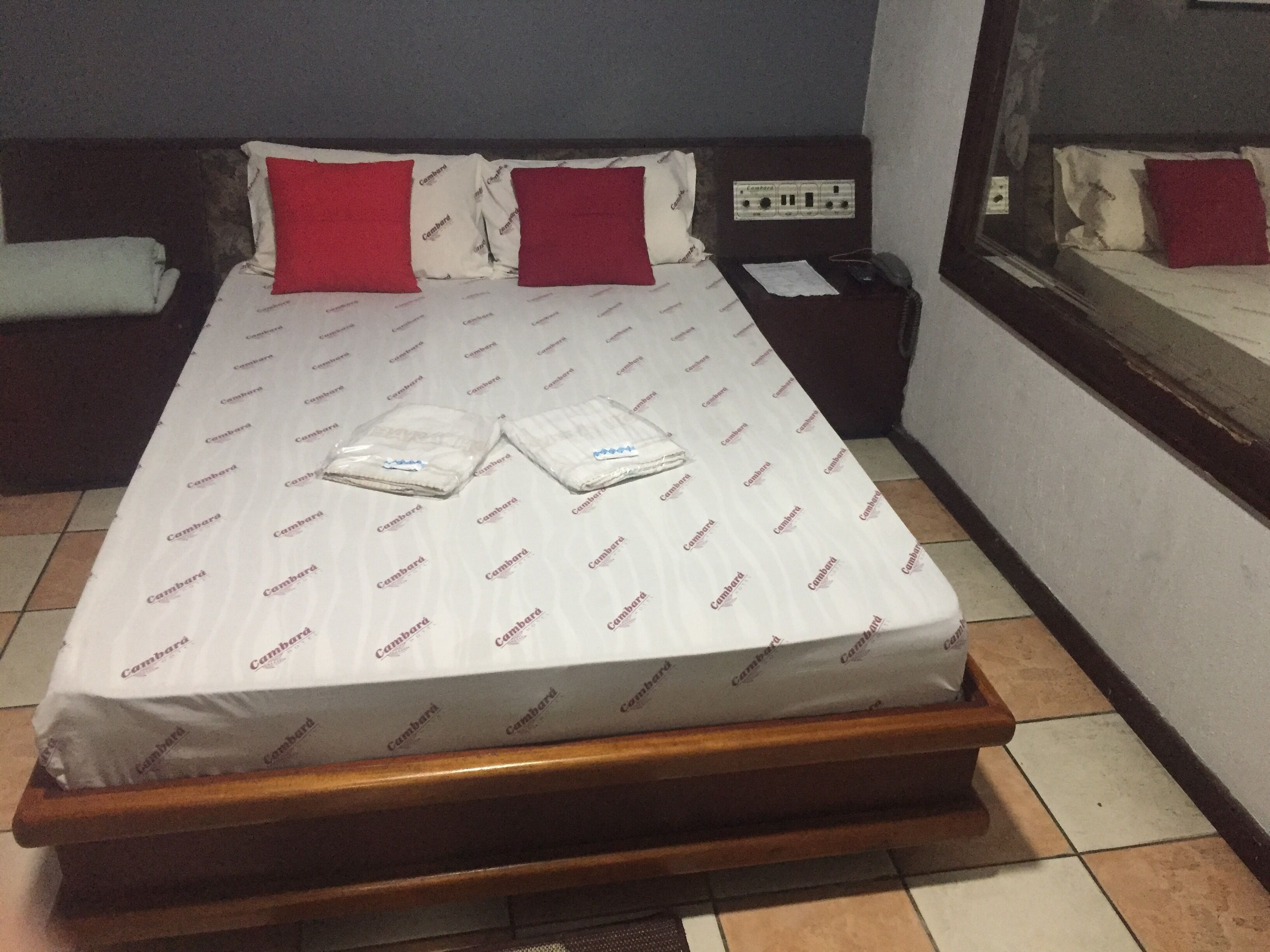 A neatly made bed with a white mattress and red pillows, a bedside table with a phone, and a window with a view of the outside.