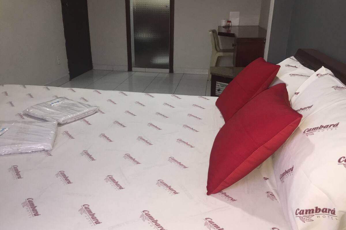 A hotel room with a bed covered in white bedding with a repeating logo, a red pillow, and a small table with a chair.