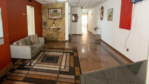 A hotel lobby with a reception desk, a sofa, a patterned rug, and a television on the wall. - Hotel Cambará (Osasco)