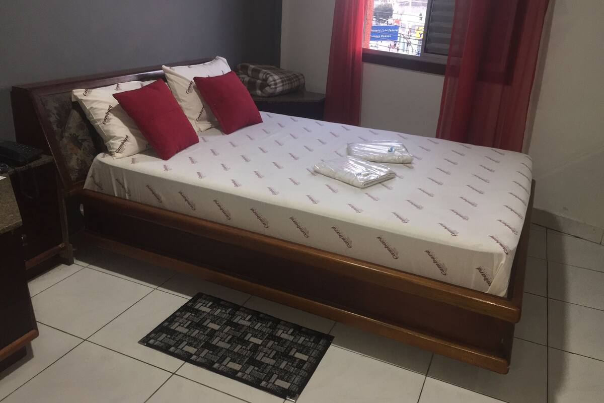 A bed with a white mattress and red pillows, a wooden headboard, and a small rug on a tiled floor.