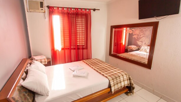 A hotel room with a bed, a TV mounted on the wall, and a window with red curtains. - Hotel Cambará (Osasco)