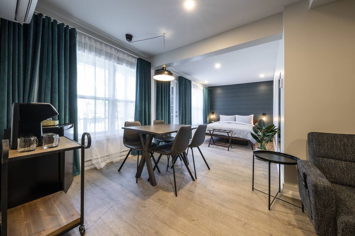 Ein modernes Hotelzimmer mit Esstisch aus Holz, Stühlen, einem kleinen Beistelltisch, einem Bett mit Kopfteil und einer Couch.