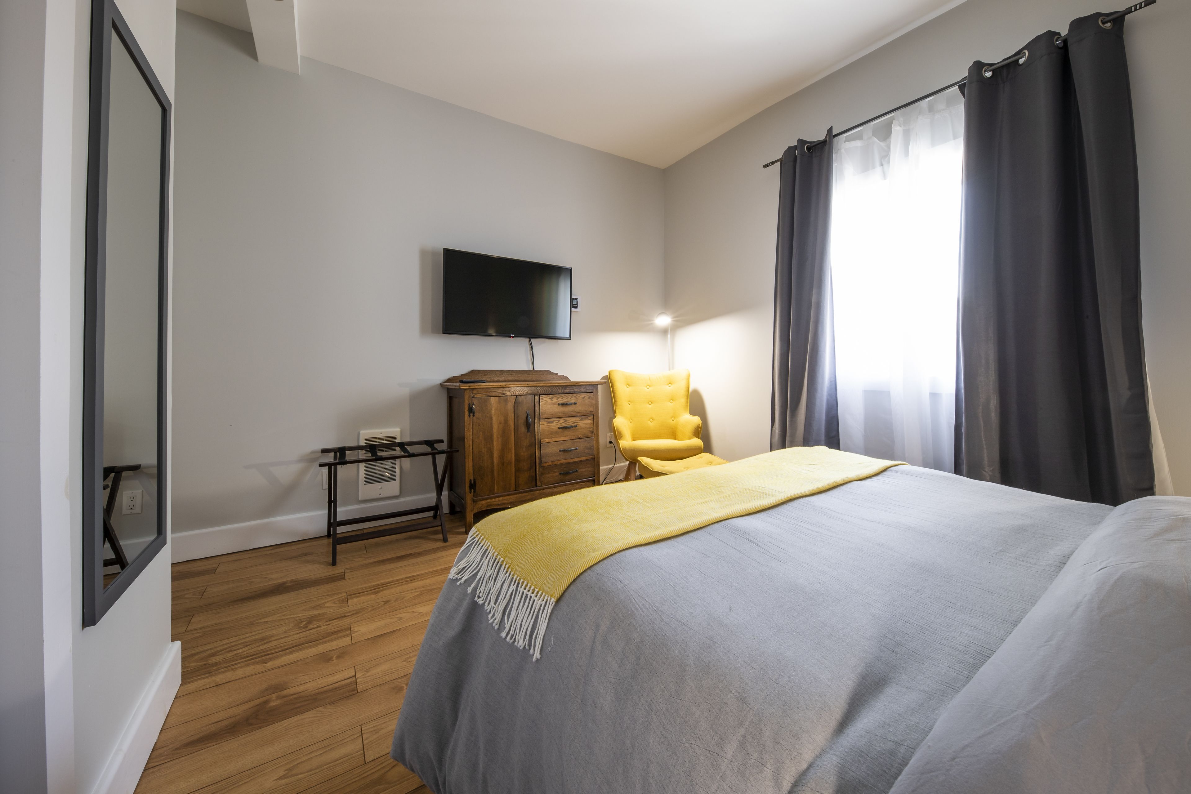 A bedroom with a bed, a wooden dresser, a yellow armchair, a flat-screen TV mounted on the wall, and a window with curtains.