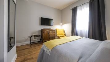 A bedroom with a bed, a wooden dresser, a yellow armchair, a flat-screen TV mounted on the wall, and a window with curtains.