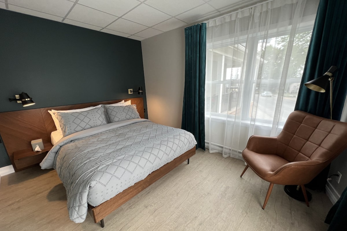 Ein Schlafzimmer mit Bett, Stuhl, Nachttisch, Lampe und einem Fenster mit Vorhängen.