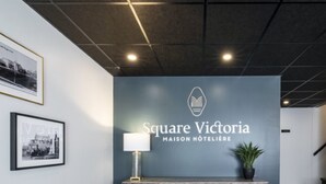 Een receptie met een bord waarop 'Square Victoria Maison Hôtelère' staat en een modern houten kastje met een lamp.
