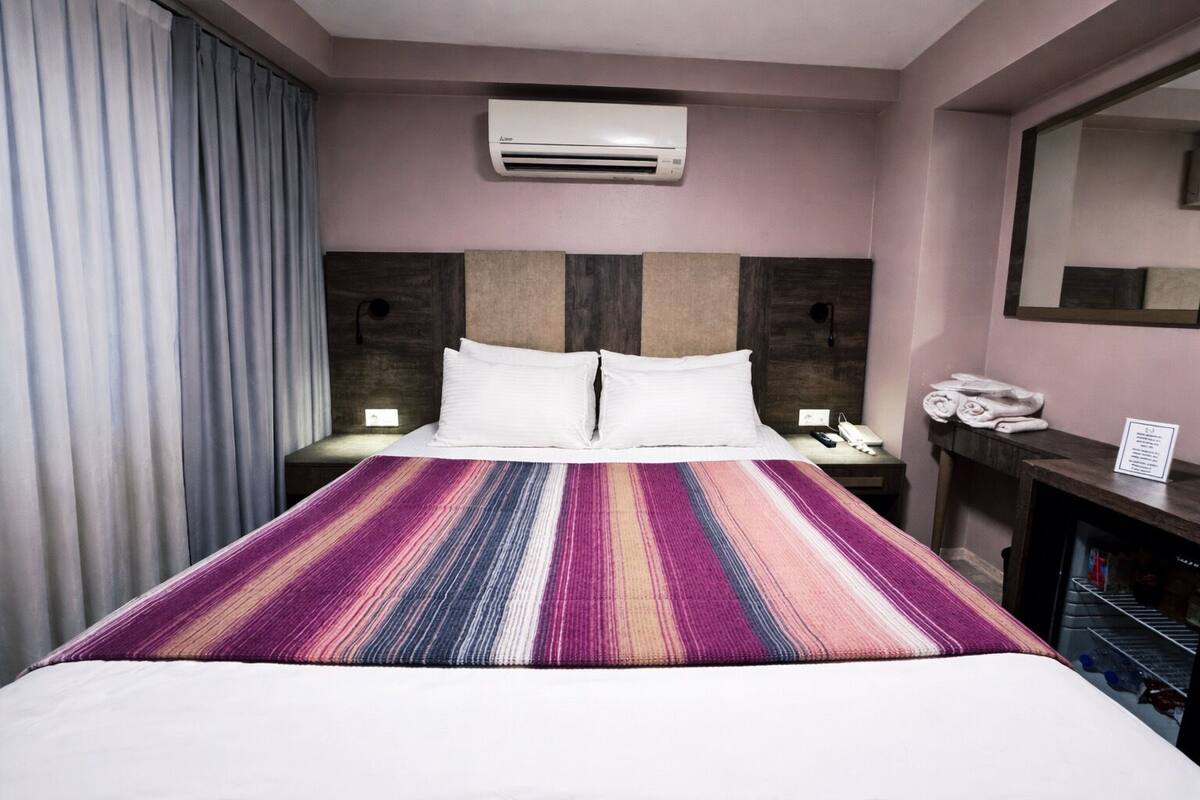 Una habitación de hotel con cama, mesita de noche, escritorio, aire acondicionado y espejo.