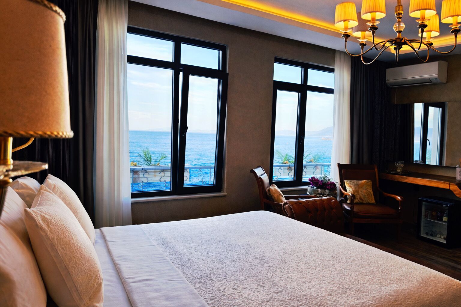 A hotel room with a large bed, two windows with a view of the sea, a desk with a chair, and a chandelier.