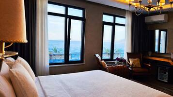 A hotel room with a large bed, two windows with a view of the sea, a desk with a chair, and a chandelier.