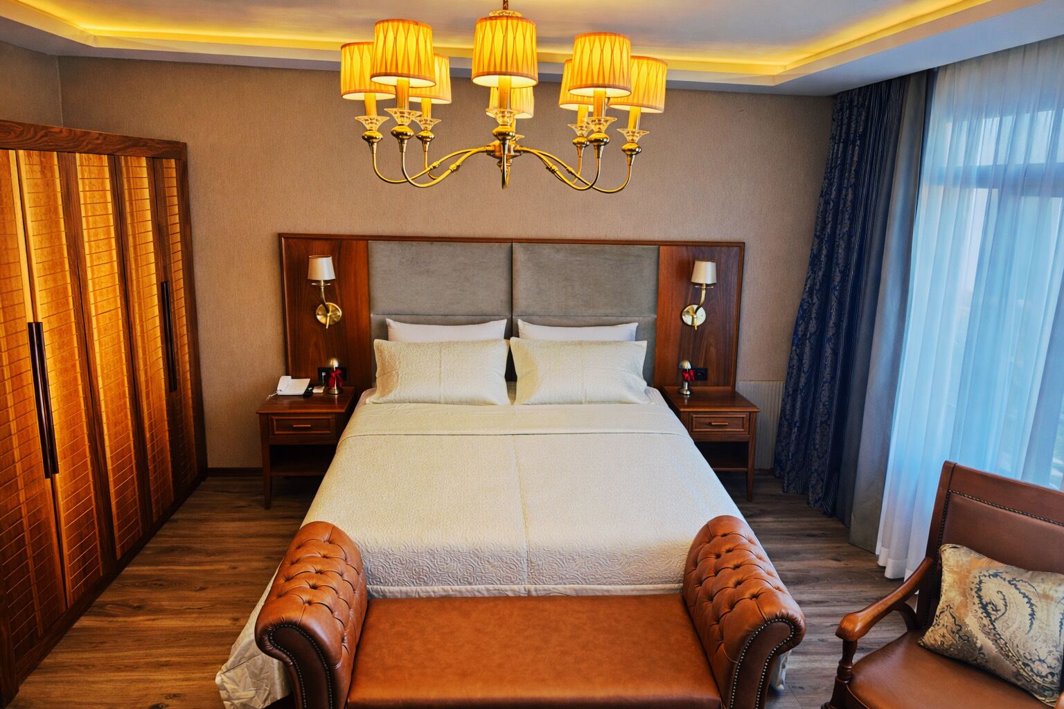 A hotel room with a large bed, wooden wardrobe, two bedside tables with lamps, a chandelier, and a view of the outside through a window with curtains.