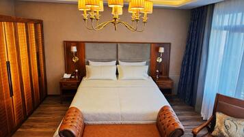 A hotel room with a large bed, wooden wardrobe, two bedside tables with lamps, a chandelier, and a view of the outside through a window with curtains.