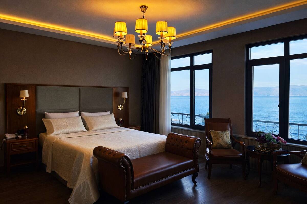 Habitación de hotel con una cama grande, cabecera de madera, candelabro, vista al mar y un espacio de descanso con una mesa y sillas.