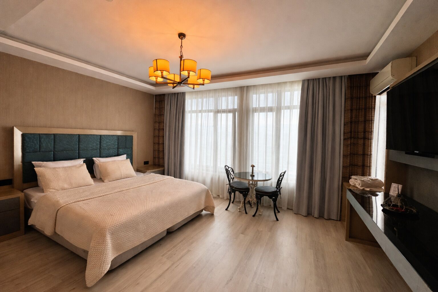 A hotel room with a large bed, a dining table with chairs, a television, and a chandelier.