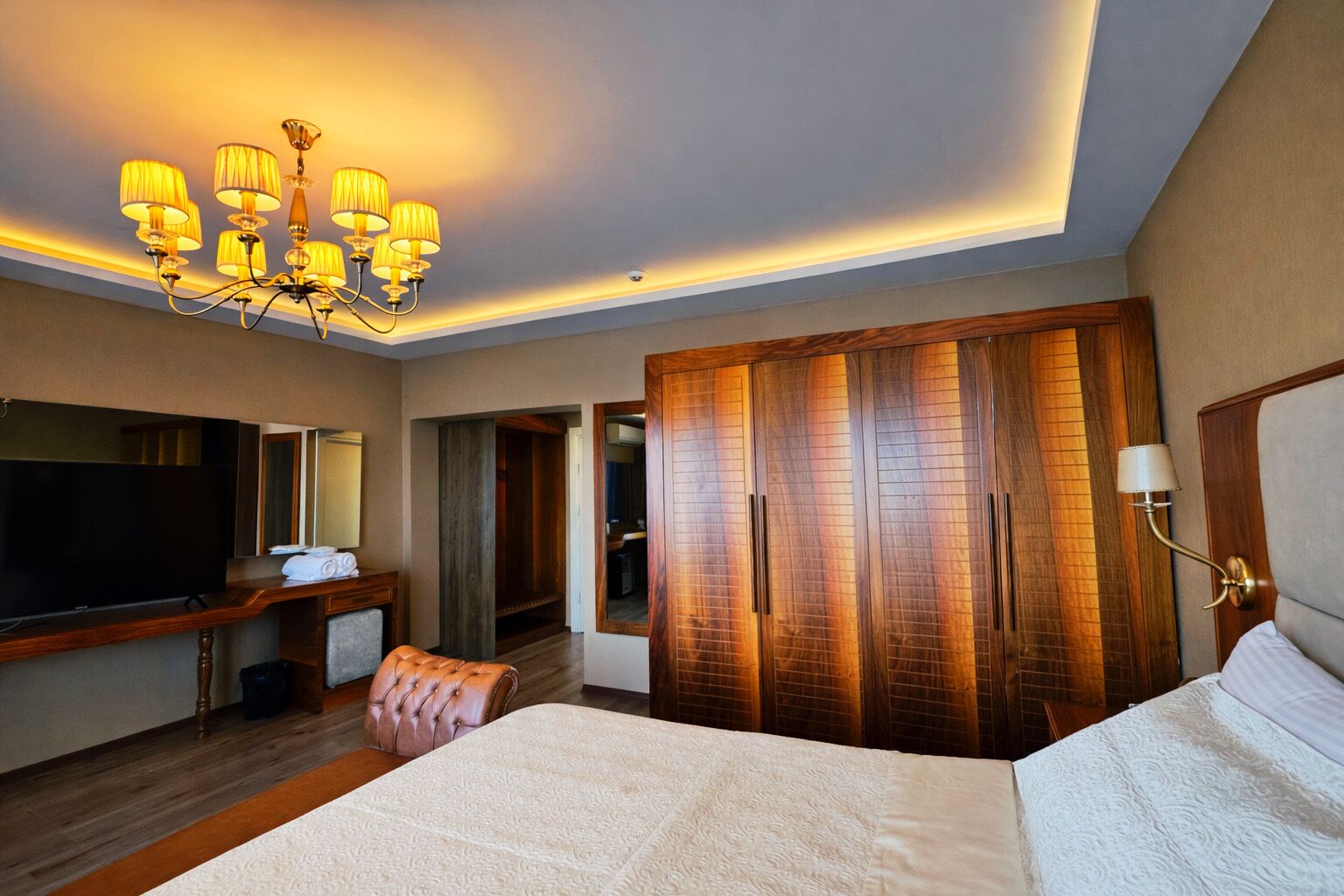 Une chambre d’hôtel avec un grand lit, une télévision, une armoire en bois, un lustre et un miroir.