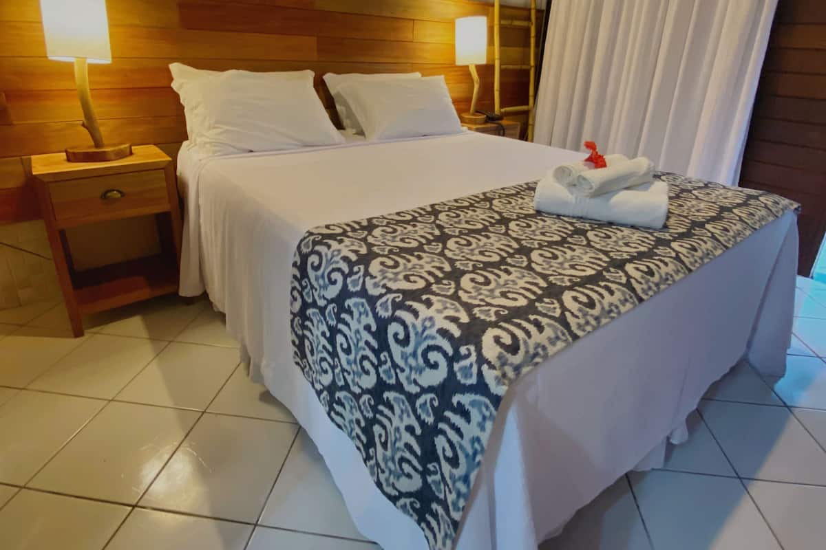 Habitación de hotel con cabecera de madera, dos lámparas de noche, cama con colcha estampada, mesita de noche y ventana con cortinas.
