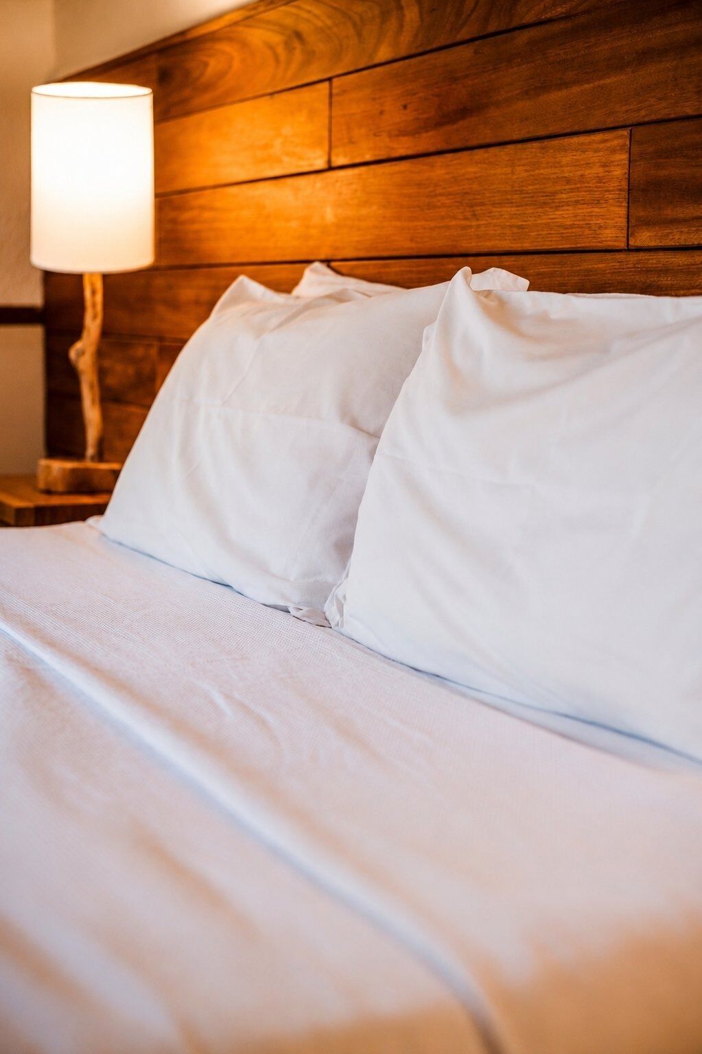 Uma cama com lençóis e almofadas brancos, uma cabeceira de madeira e uma mesa de cabeceira com abajur.