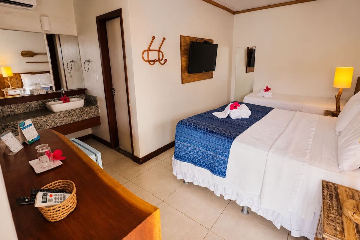 A hotel room with a bed, bedside table, lamp, and a small table with a remote control and a basket.
