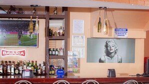 A bar area with a Bavaria sign, a large TV screen, various bottles, and a framed picture on the wall. - Hotel Arrecifes (San José)