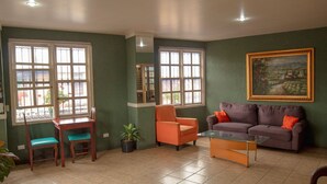 A living room with a green wall, a purple sofa, an orange armchair, a wooden table with a teal chair, and a painting. - Hotel Arrecifes (San José)