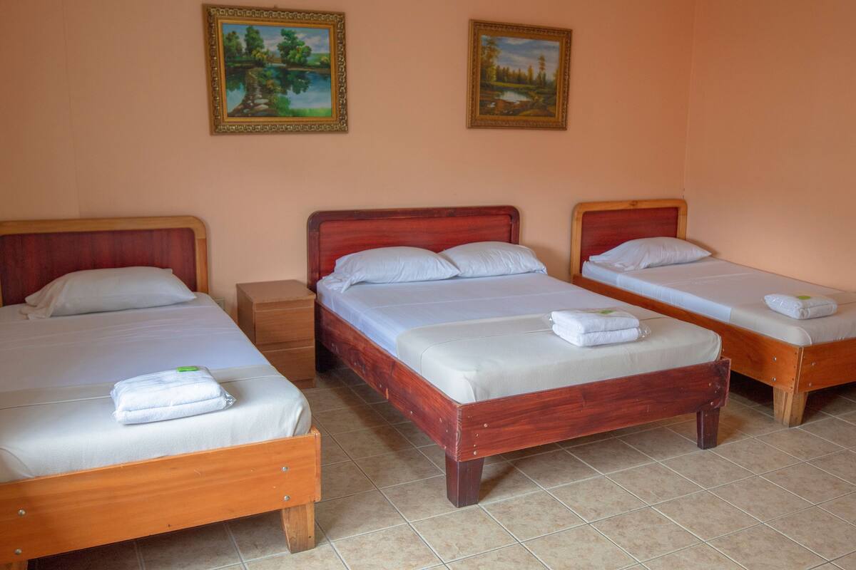 A hotel room with three wooden beds, each with white linens and pillows, and two framed landscape paintings on the wall.