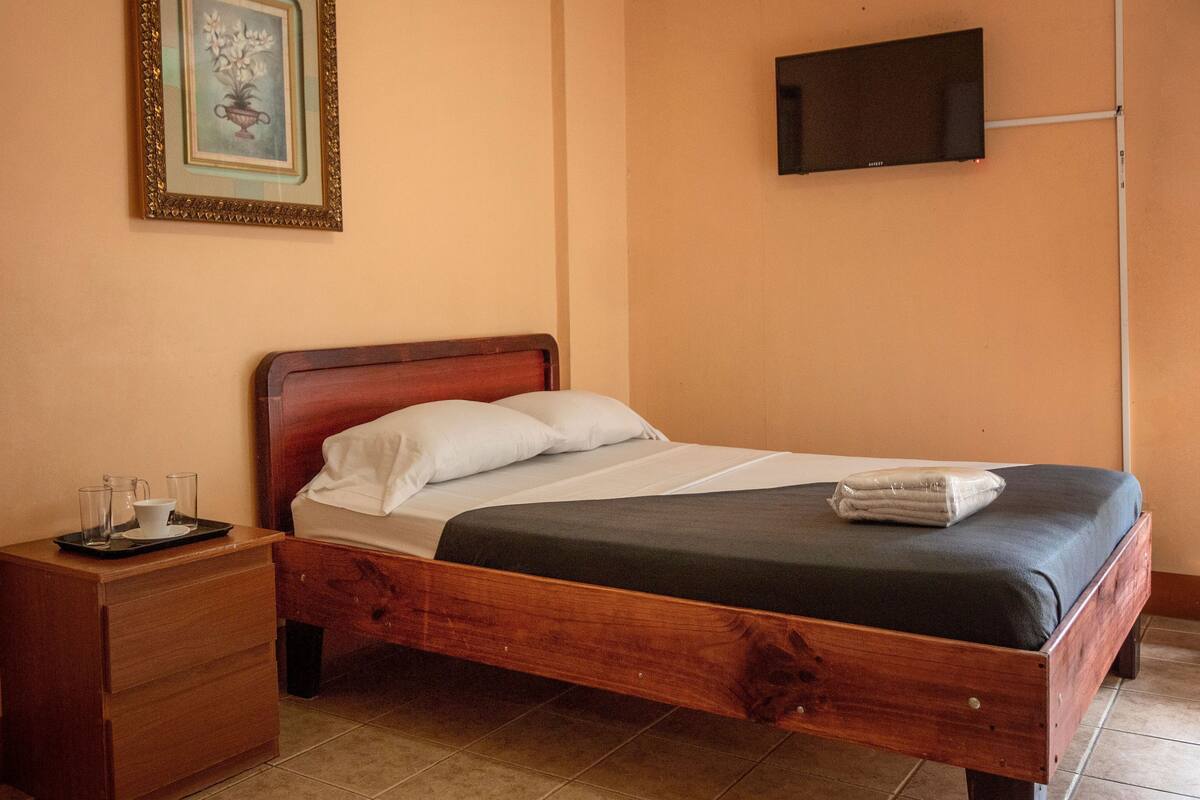 A wooden bed with white linens, a small wooden nightstand with a tray of glasses, and a framed picture on the wall.