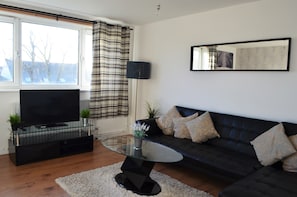 A modern living room with a glass coffee table, a black sofa, a flat-screen TV, and a large mirror on the wall.