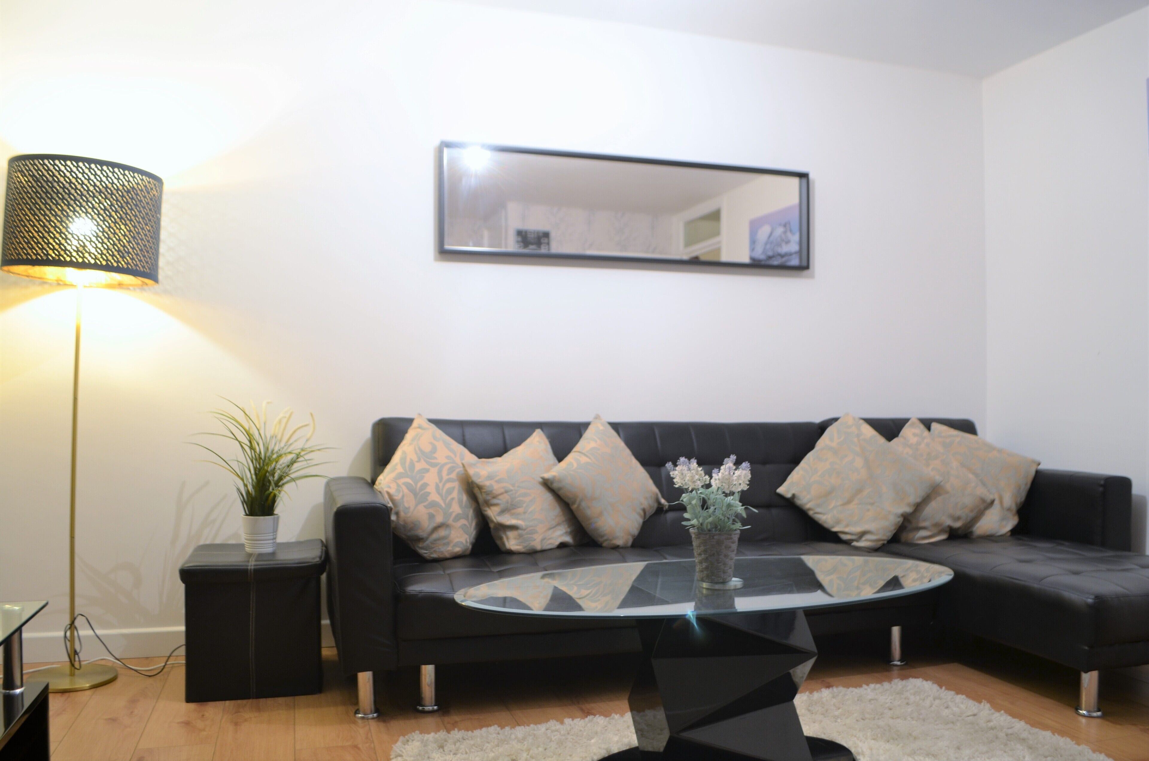 A modern living room with a black sofa, a glass coffee table, a floor lamp, and a mirror on the wall.