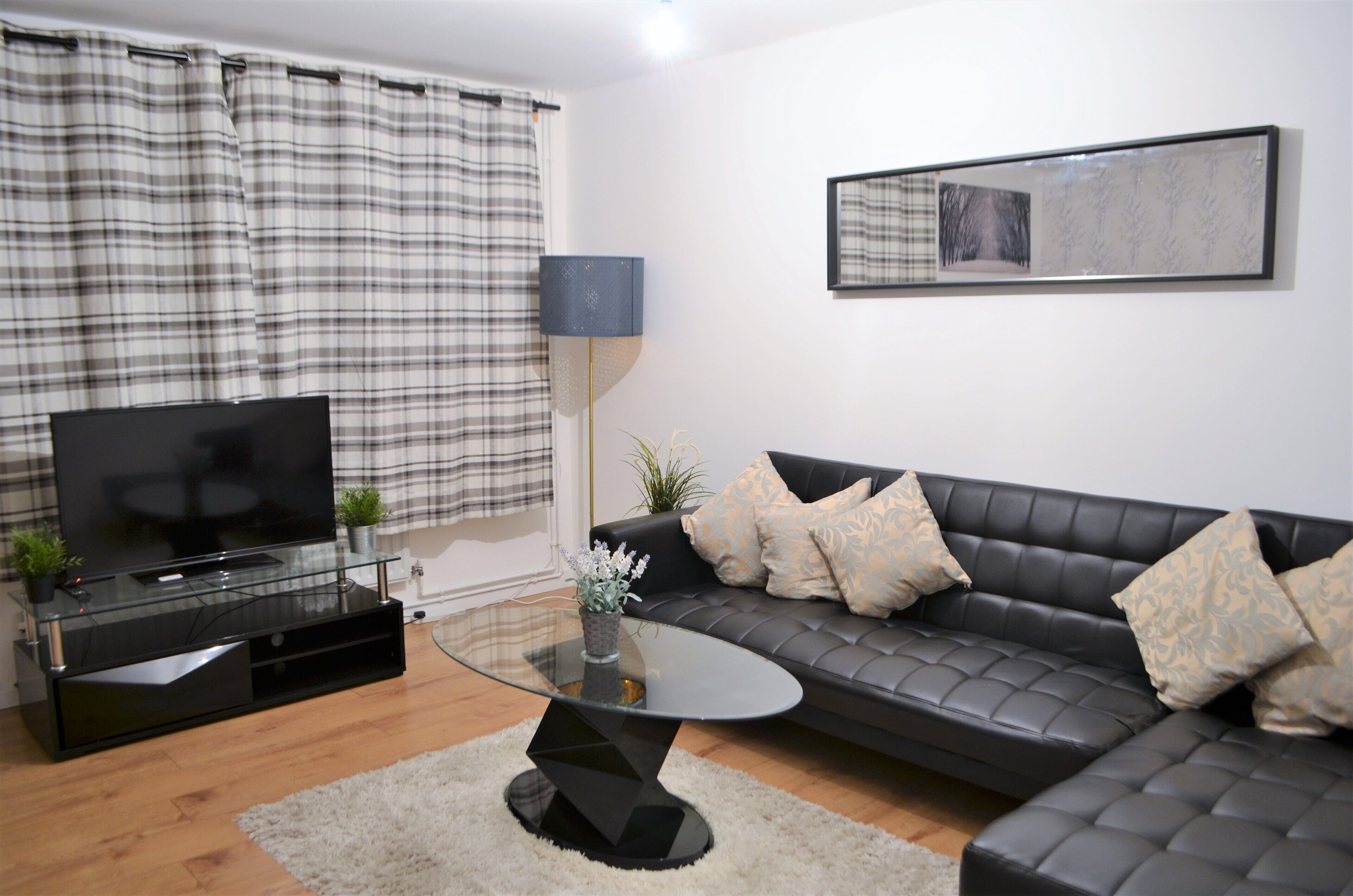 Ein modernes Wohnzimmer mit einem schwarzen Ledersofa, einem Glastisch, einem Flachbildfernseher auf einem schwarzen Standfuß und einem karierten Vorhang.