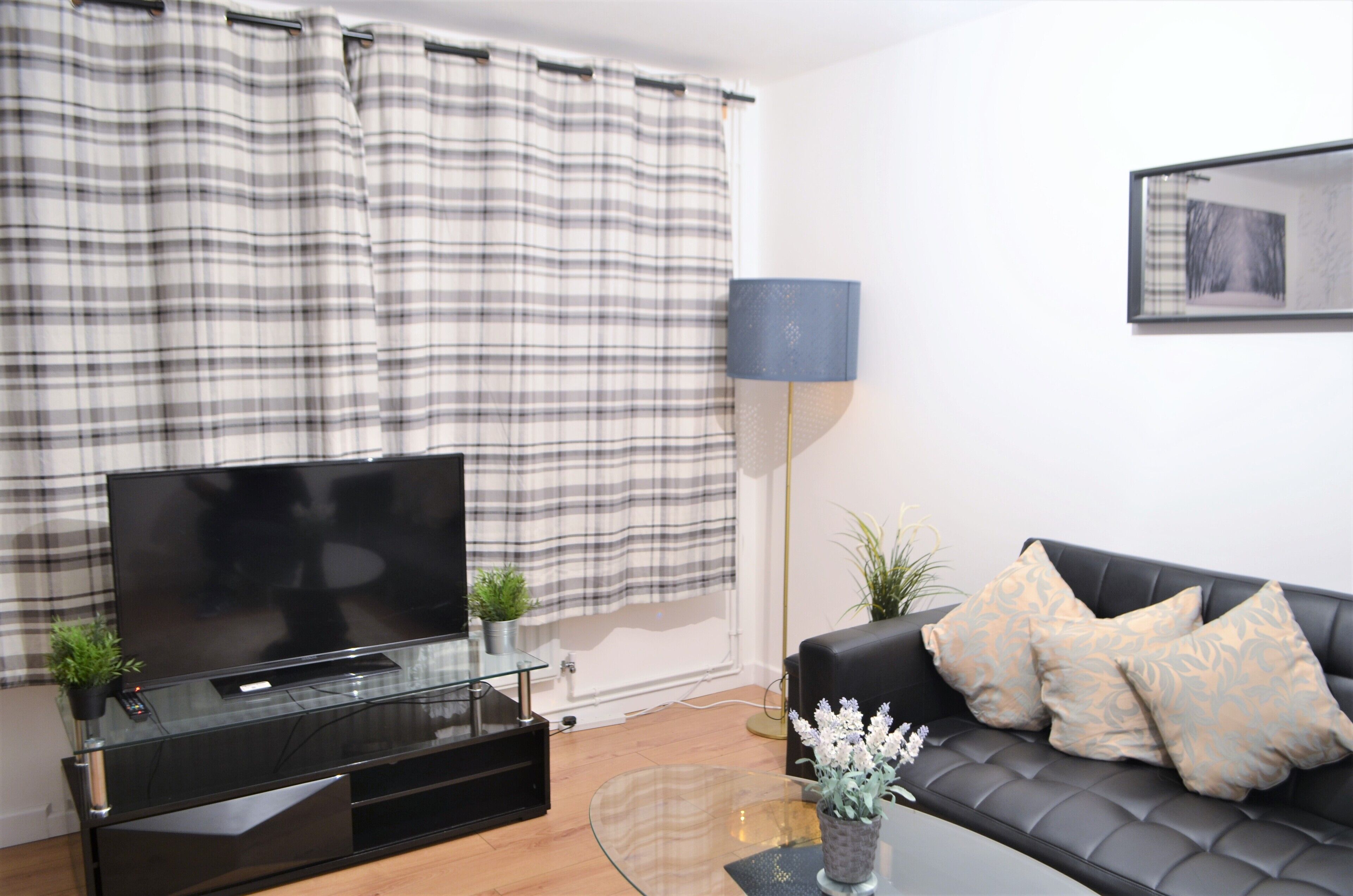 Ein modernes Wohnzimmer mit Flachbildfernseher auf einem Glastisch, einer schwarzen Ledersofa mit gemusterten Kissen, einer Stehlampe und einem großen Fenster mit Karottenvorhängen.