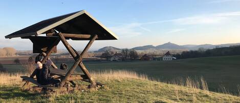 Een houten gebouw met een schuin dak in een grasveld, omringd door huizen en heuvels op de achtergrond.