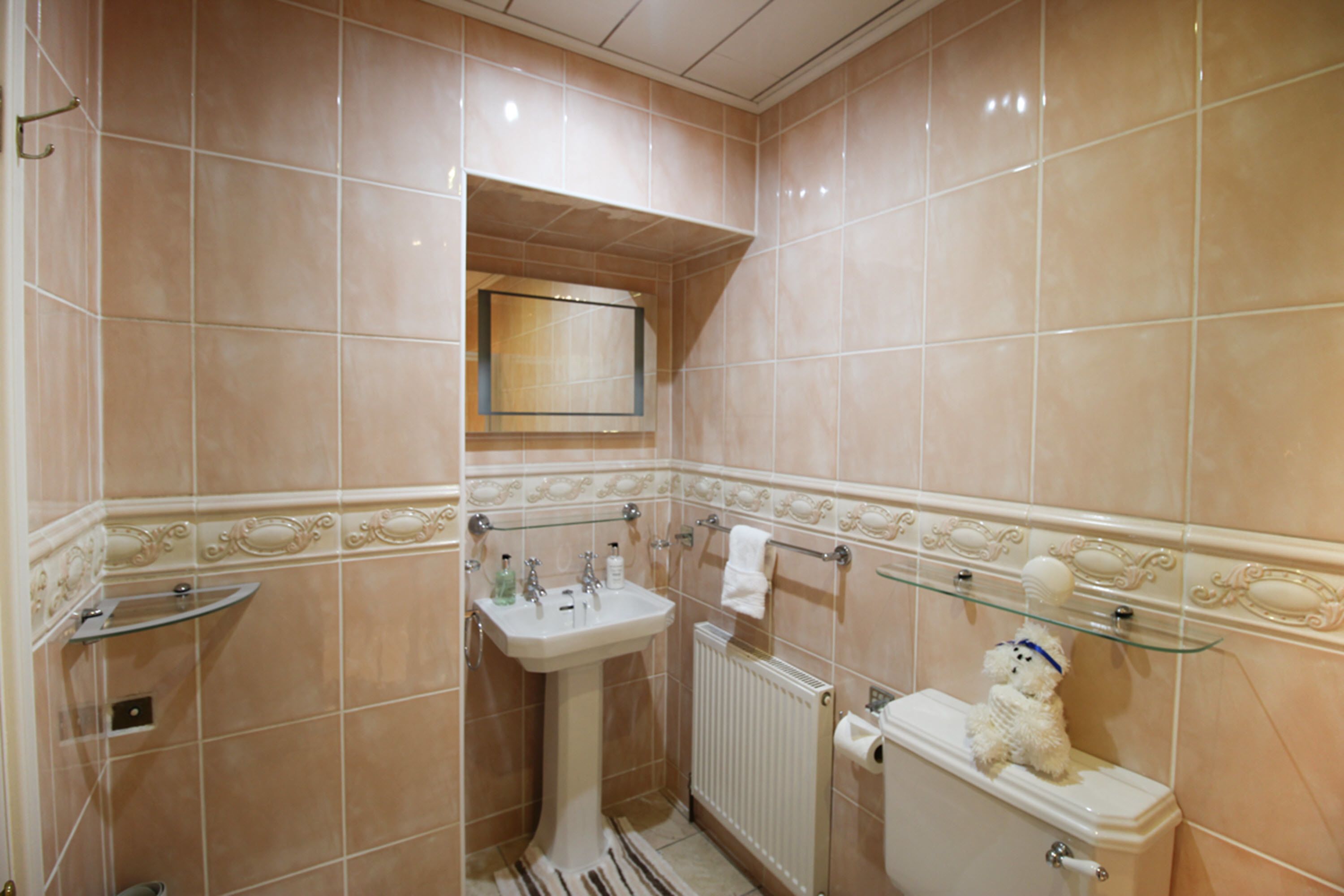 A bathroom with beige tiles, a pedestal sink, a toilet, a glass shelf, and a towel rack.