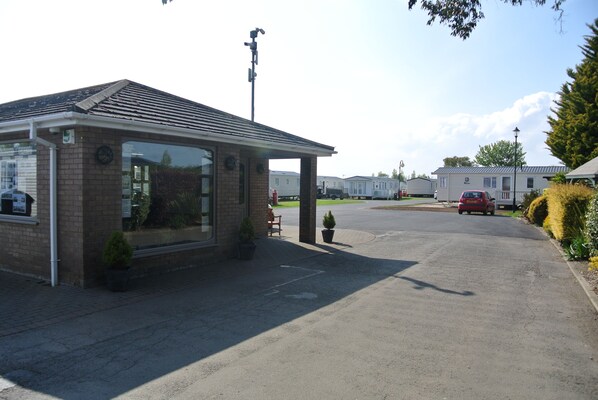 A small brick building with a covered entrance, a parking area, and mobile homes in the background.