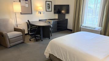 Hotellihuone, jossa on sänky, työpöytä, tuoli, televisio, lamppu, ilmoitustaulu ja ikkuna, jossa on verhot.