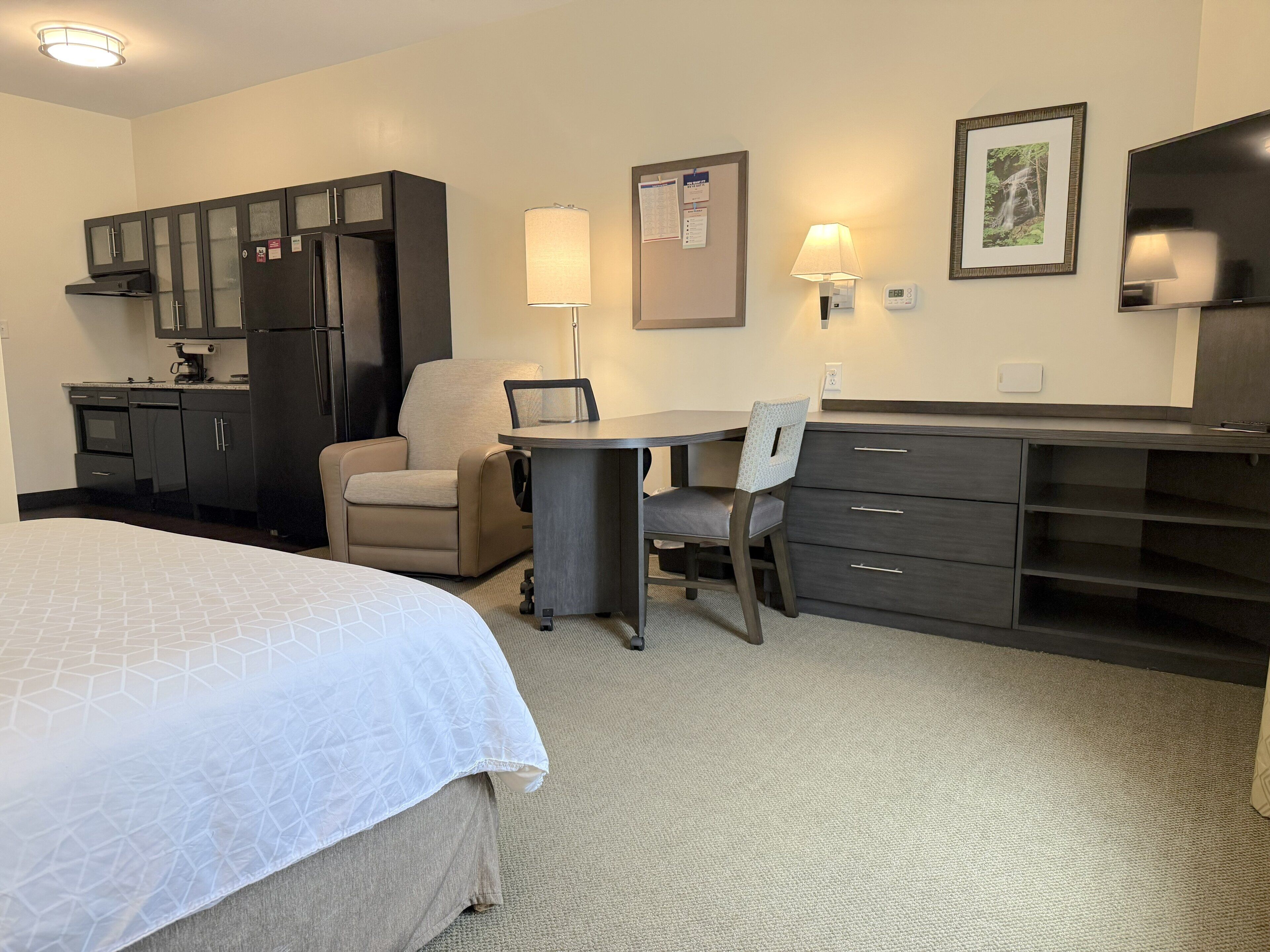 A hotel room with a bed, a desk, a chair, a kitchenette, and a TV.