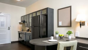 A compact kitchen with dark cabinetry, a built-in refrigerator, a dining table with a chair, and a wall-mounted lamp. - Candlewood Suites Jacksonville - Mayport by IHG (Atlantic Beach)
