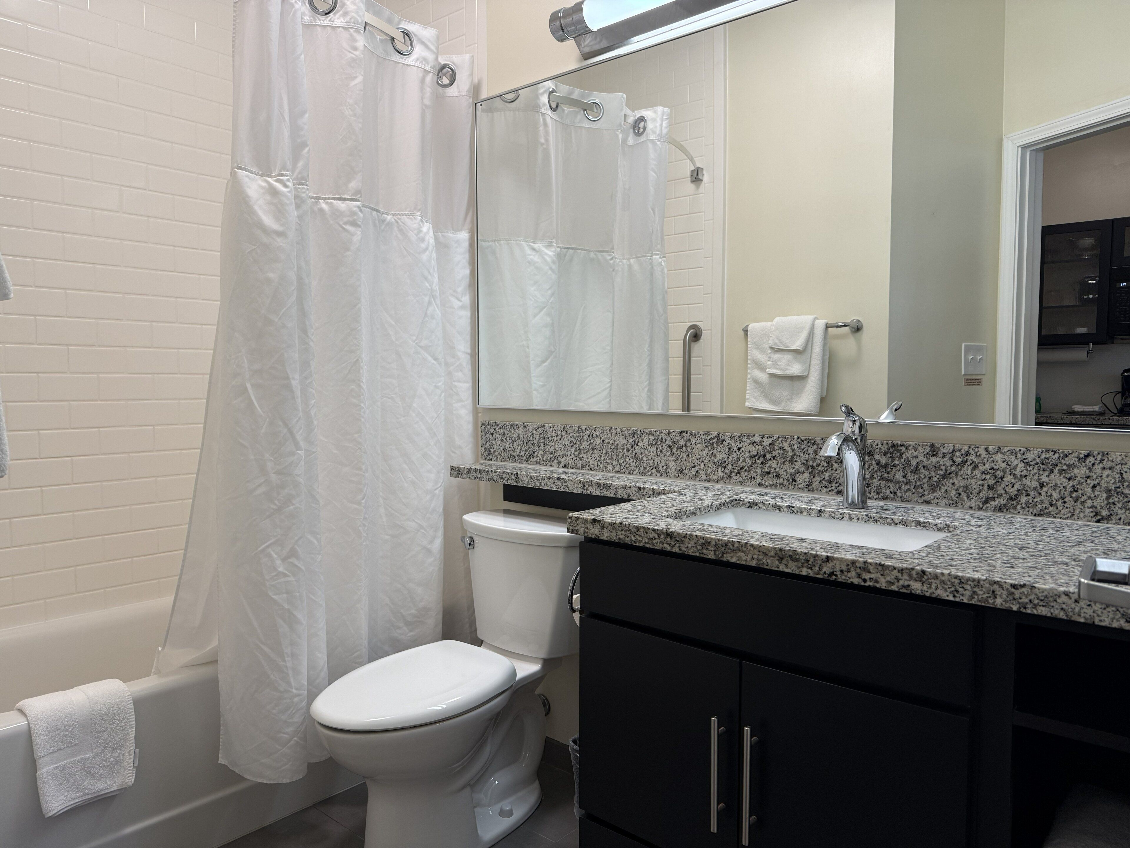 Un baño con inodoro blanco, cortina de ducha, lavabo con encimera de granito y un espejo sobre el lavabo.