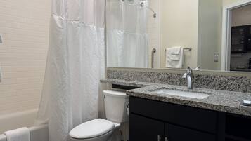 Un baño con inodoro blanco, cortina de ducha, lavabo con encimera de granito y un espejo sobre el lavabo.