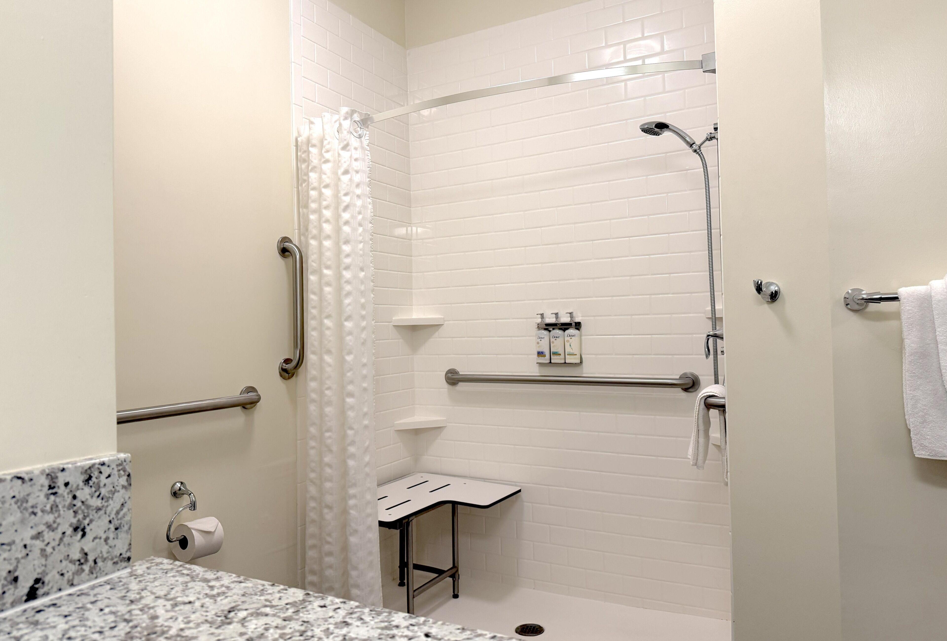 A bathroom with white tiled walls, a shower with a handheld showerhead, a grab bar, a towel rack, and a marble countertop.