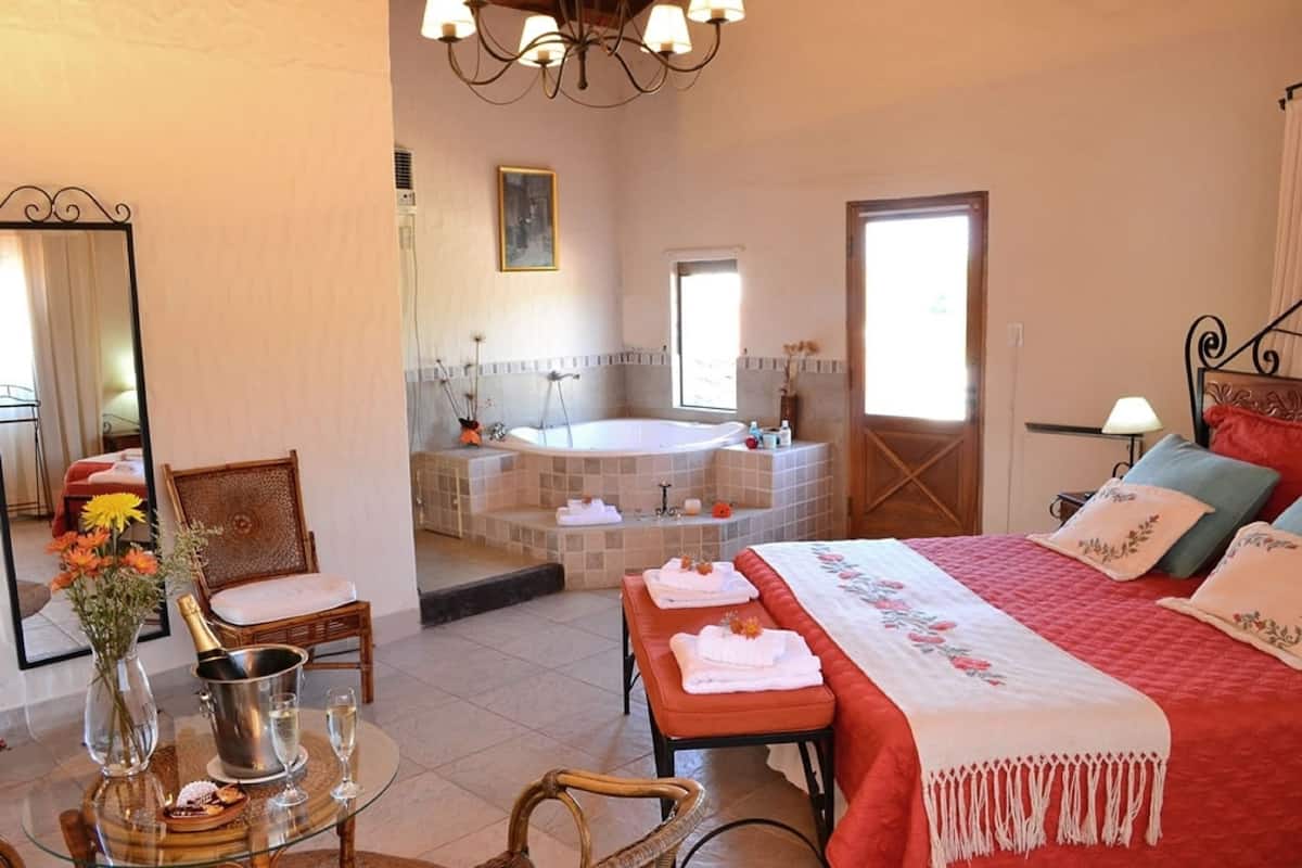A spacious bedroom with a large bed, a sitting area with a chair and a small table, a bathtub, and a large mirror.