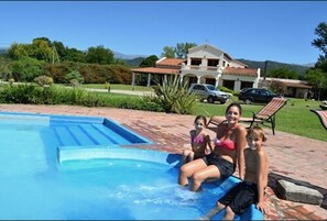A swimming pool with a clear blue water feature, surrounded by a brick patio and a large house with a tiled roof in the background. - Hosteria La Caldera (La Caldera)